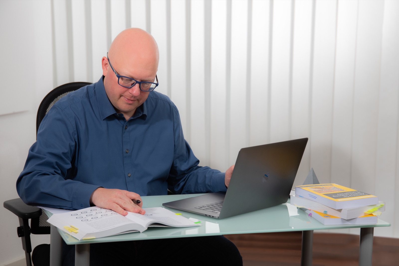 Thorsten Jungmann, ein Mann mit kurzen Haaren, dunkler Brille und dunkelblauem Hemd, sitzt am Schreibtisch vor einem Computer und liest in einem Buch.